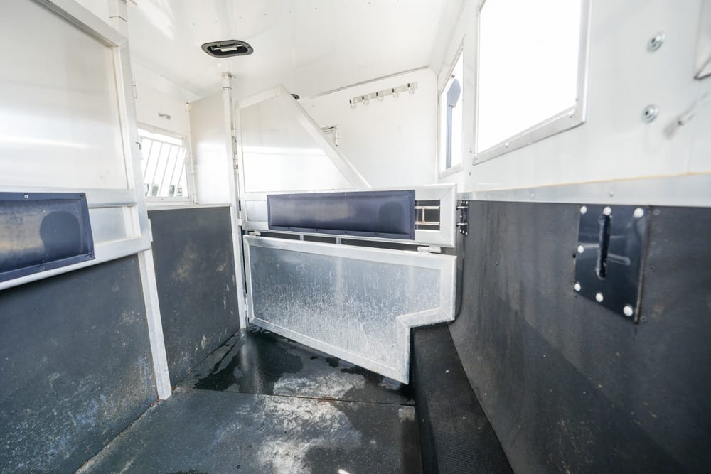 Interior of a white horse trailer with a dividing metal stall, dark rubber flooring, and dusty walls at the back of the space.