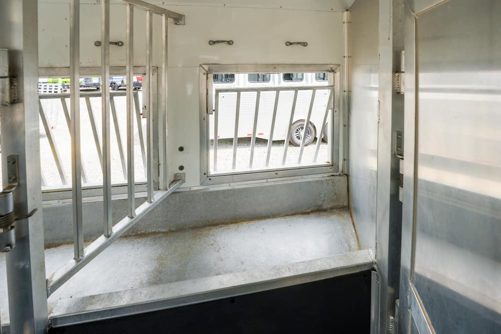 Interior of a metal livestock trailer with vertical bars and a small window looking out to parked cars. A grated partition divides the space.