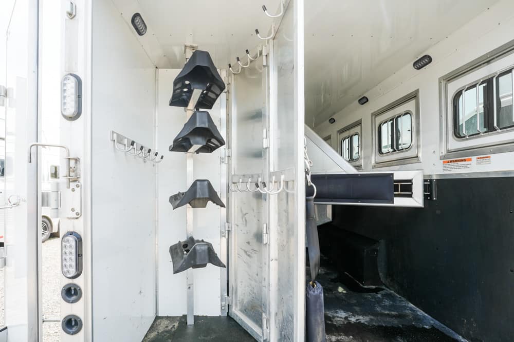 Interior of a horse trailer showing a vertical rack with four black saddle cradles and multiple wall hooks, separating stall area from the living area with windows along the side.