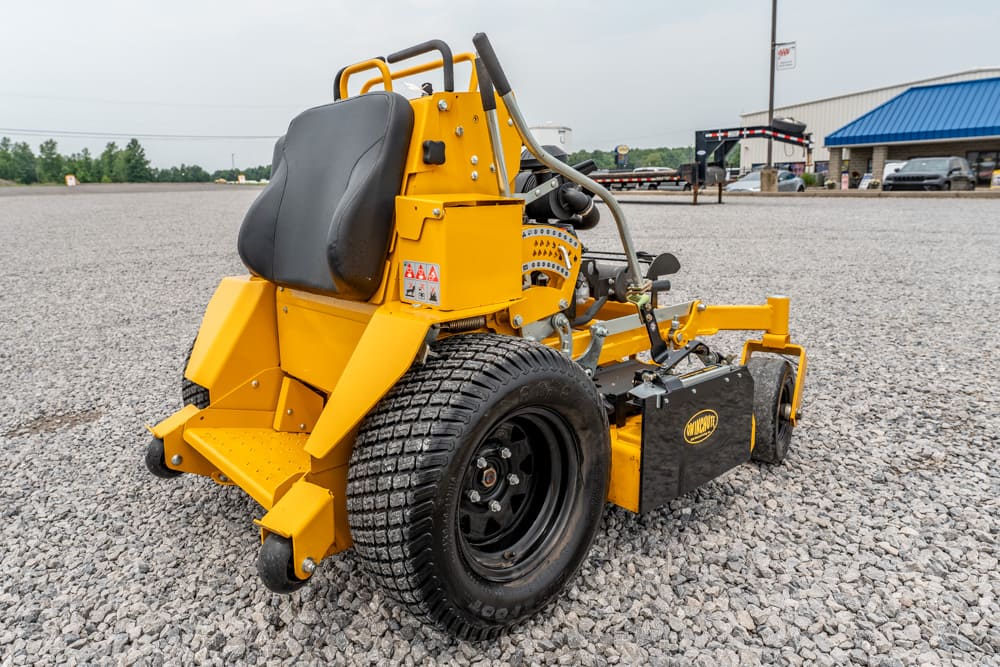 Yellow commercial walk-behind lawn mower with a black seat and controls, parked on a gravel lot by a building.