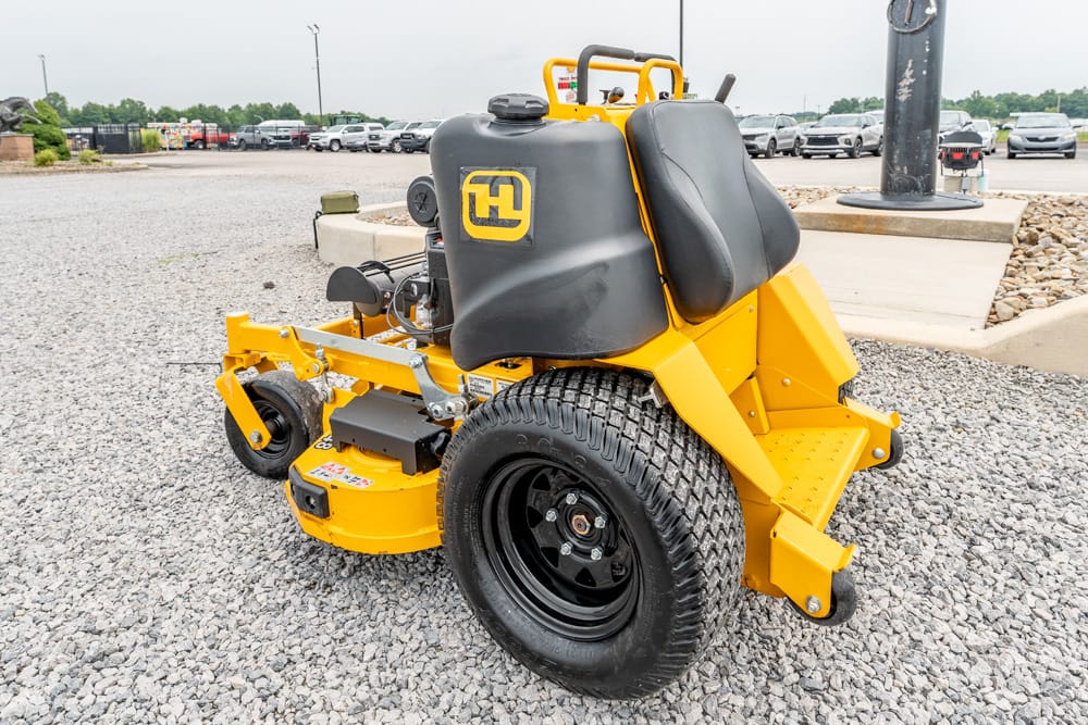 Yellow commercial riding mower with a black seat and large rear wheel, parked on a gravel lot.