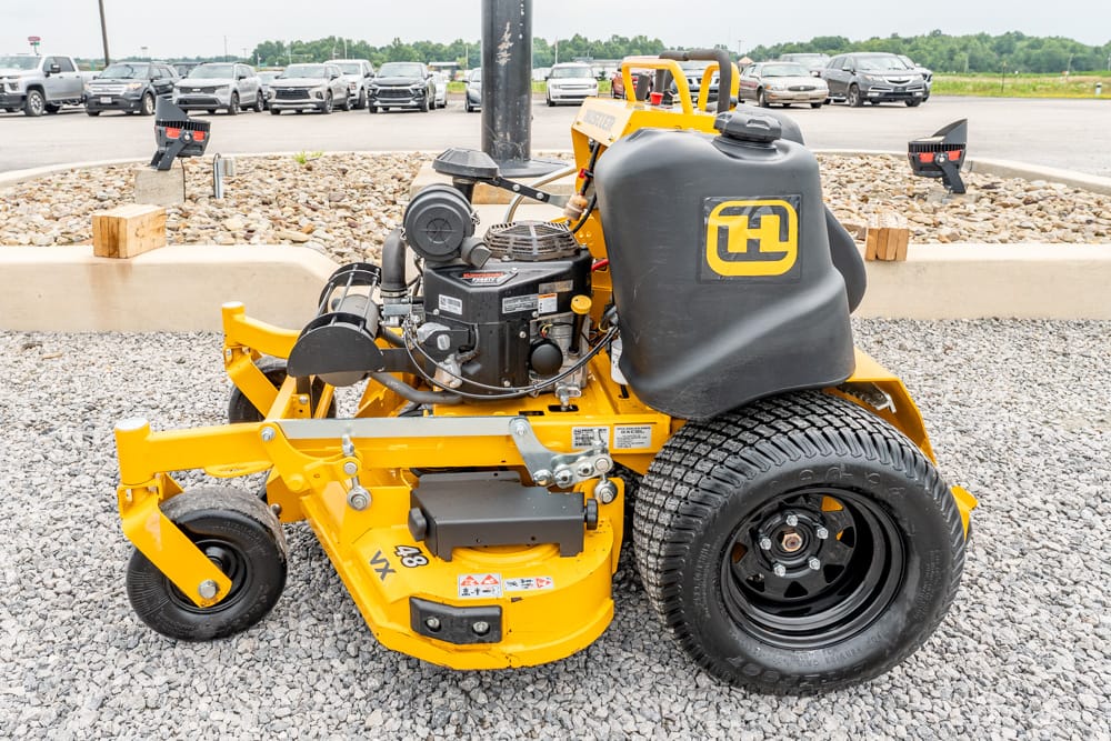 Yellow zero-turn riding lawn mower with a black seat and large rear tire, parked on a gravel lot near a row of cars in the background (brand logo on seat).