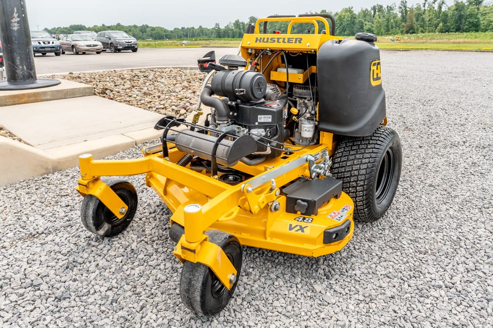 Yellow Hustler zero-turn lawn mower with exposed engine on a gravel lot, large rear tire and forward caster wheels (48 in deck)