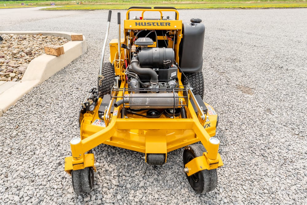 Front view of a yellow Hustler turf machine with exposed engine on a gravel parking area.
