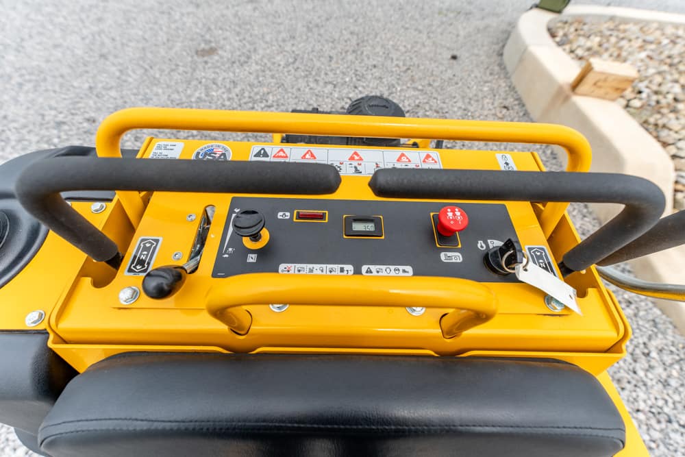 Close-up of a yellow industrial control panel with two black handrails, joysticks, and a red emergency stop button surrounded by warning labels.