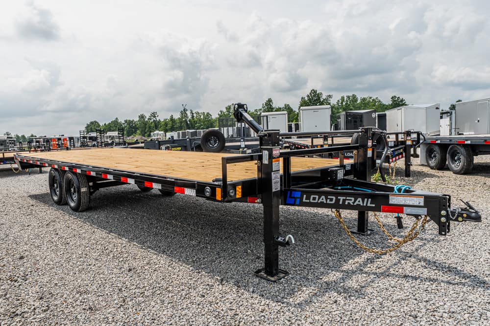 Flatbed trailer with a wooden deck and dual axles in a gravel trailer lot; brand label 'Load Trail' on the tongue, surrounded by other trailers.