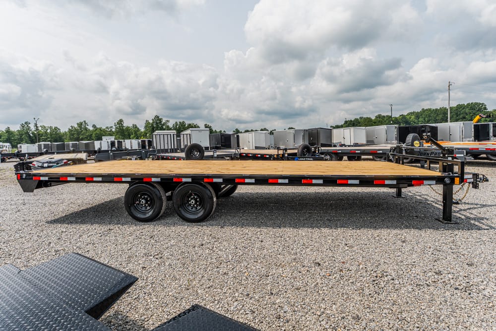 Long flatbed trailer with a wooden deck and dual rear wheels parked on a gravel lot, with rows of trailers in the background.