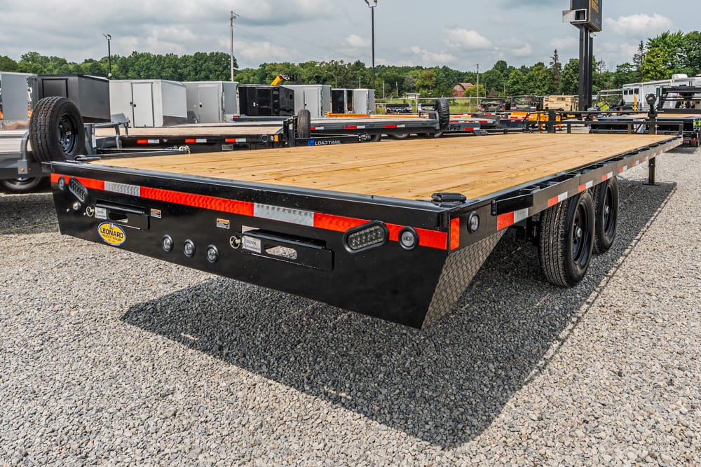 Flatbed trailer with a wooden deck and black steel frame in a gravel lot, showing multiple axles and rear lights.
