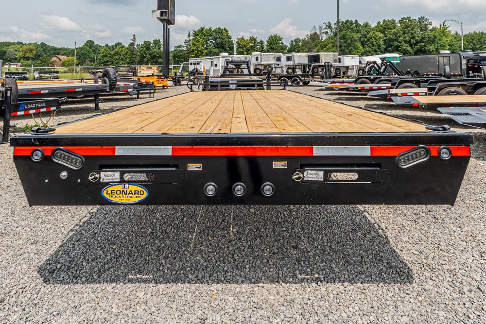 Flatbed trailer with wooden deck seen from the rear, parked on a gravel lot with other trailers in the background.