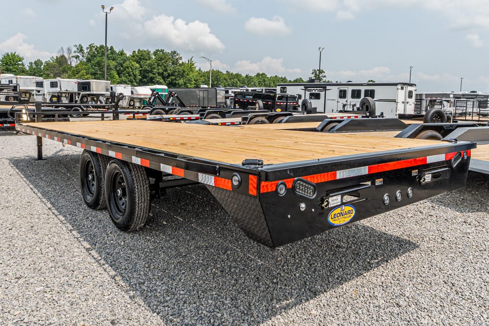 Flatbed trailer with a wooden deck and dual-axle wheels on a gravel lot, with other trailers in the background.