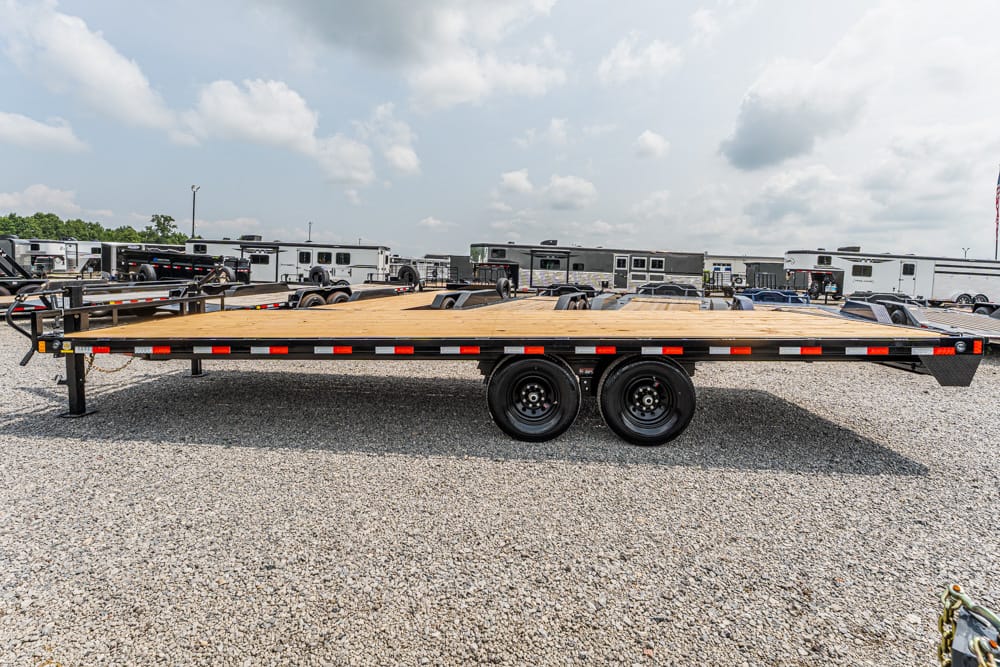 Flatbed trailer with a wooden deck and dual rear wheels parked in a lot of RV and equipment trailers on gravel under a cloudy sky and a distant horizon.