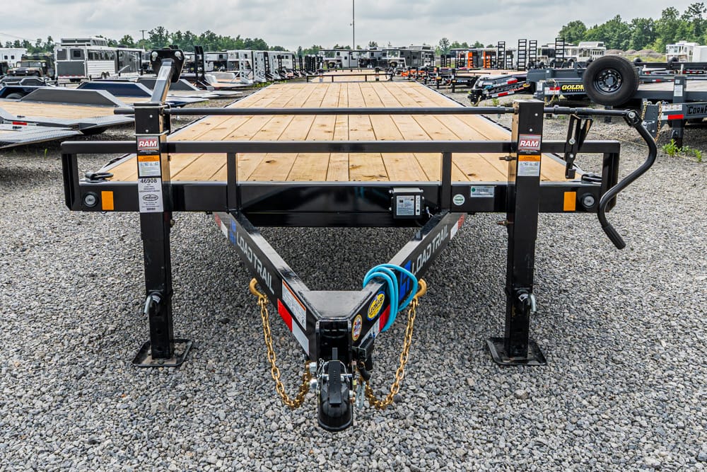 Black steel flatbed trailer with a wooden deck, safety chains and hoses at the hitch, parked on a gravel lot with other trailers in the background.