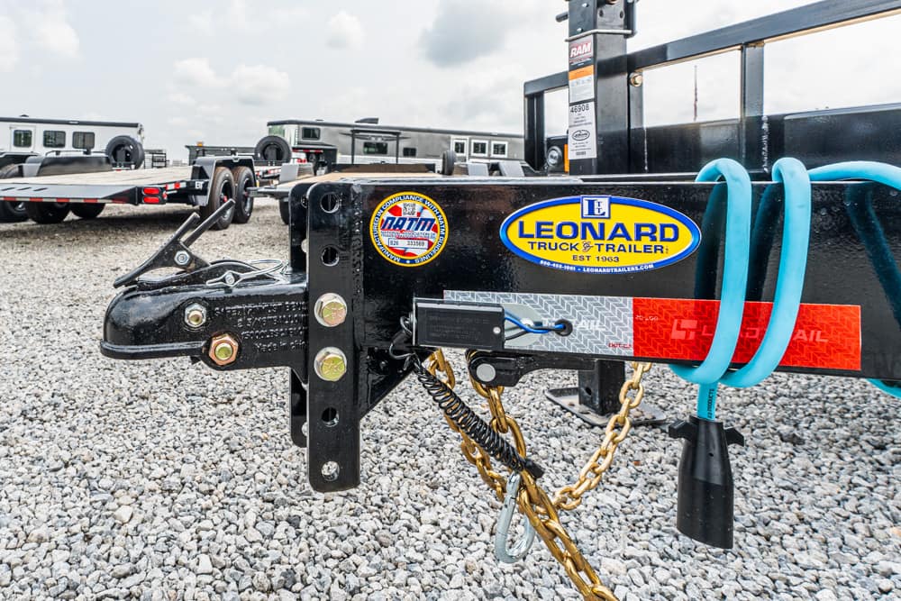 Close-up of a black trailer hitch with a Leonard Truck & Trailer sticker, blue hoses, and hanging chains on a gravel lot.