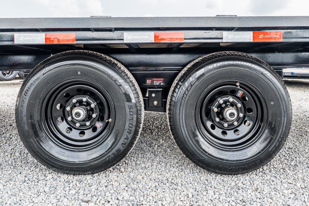 Dual trailer wheels on a black flatbed trailer, resting on gravel ground.