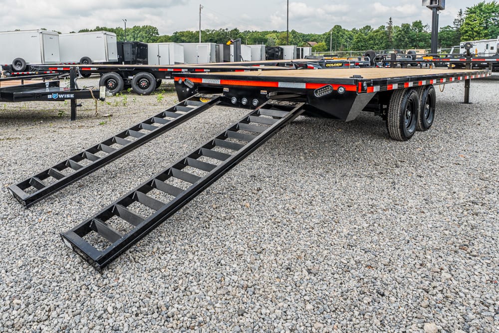 Black car-hauler flatbed trailer with two unfolded rear loading ramps on a gravel lot, with other trailers in the background.