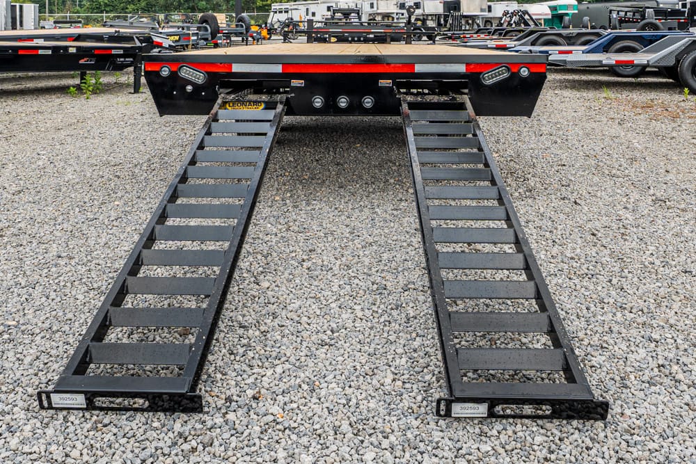 Two black loading ramps extended from a flatbed trailer onto a gravel lot, ready for vehicle loading or unloading.