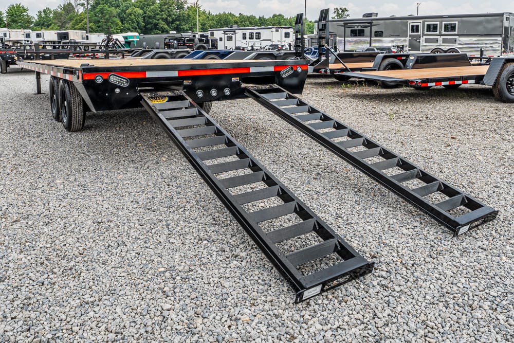 Black metal loading ramps extend from a flatbed trailer onto a gravel lot, with other trailers and trucks visible in the background, ready for loading equipment or vehicles.