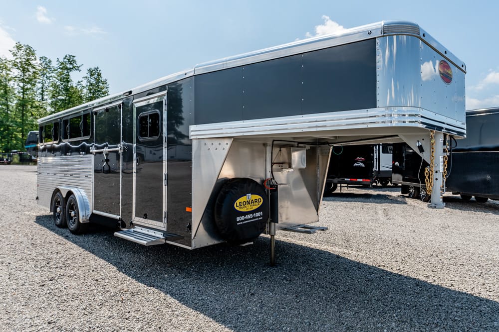 Black, multi-axle gooseneck horse trailer on a gravel lot with a spare tire at the front and trees in the background