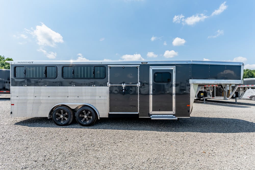 Long two-axle horse trailer with dark gray upper panels and aluminum lower siding parked on a gravel lot, with a side entry door and windows along the side.