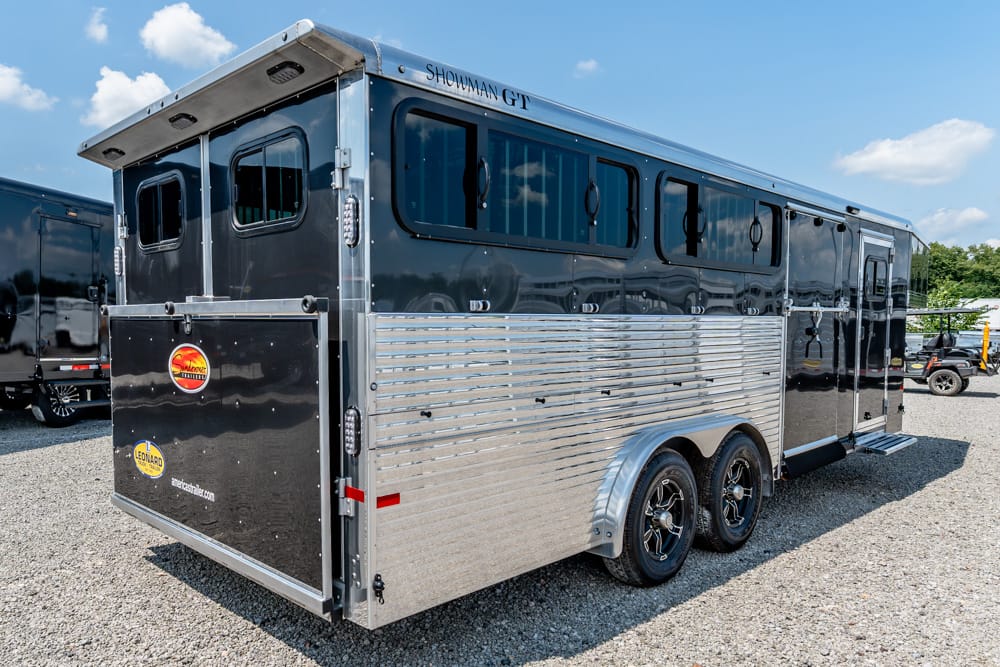 Large black and silver horse trailer with two axles parked on gravel, showing windows and a rear ramp area.