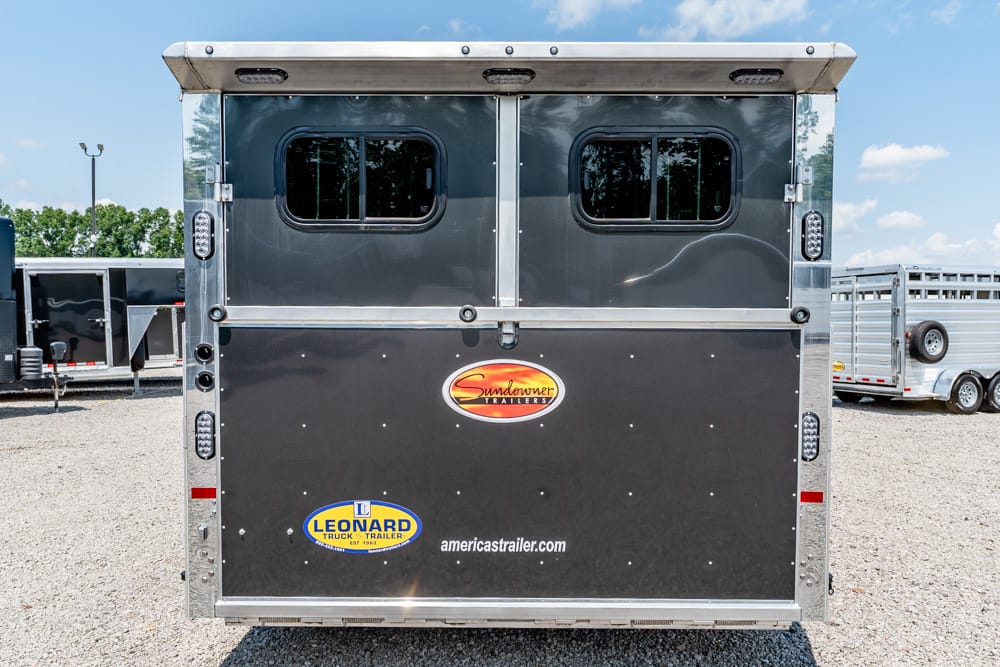 Back of a dark gray Sundowner trailer with two small windows, chrome trim, and brand stickers on a gravel lot.