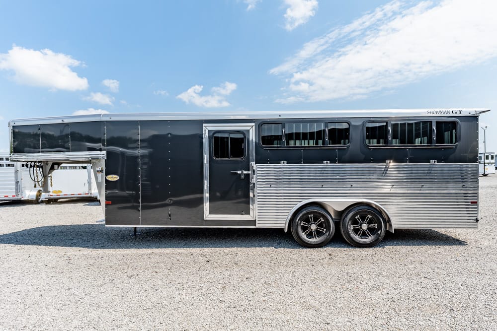 Black and silver horse trailer with two axles and windows along the top, parked on a gravel lot under a blue sky.