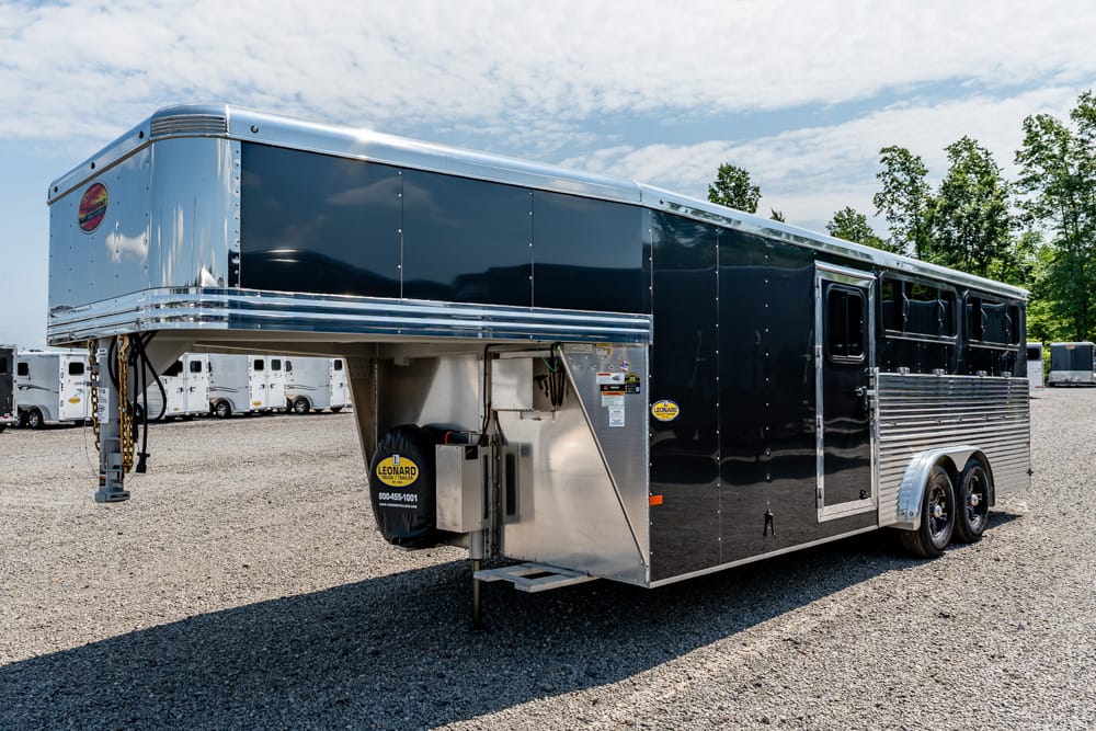 Glossy black gooseneck horse trailer with chrome trim and dual axles, parked on a gravel lot with other trailers nearby.
