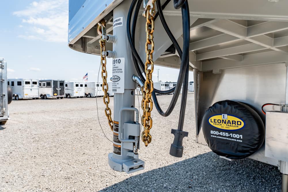 Close-up of a trailer hitch with gold safety chains and hoses, spare tire labeled Leonard Trailer in a row of parked trailers behind.