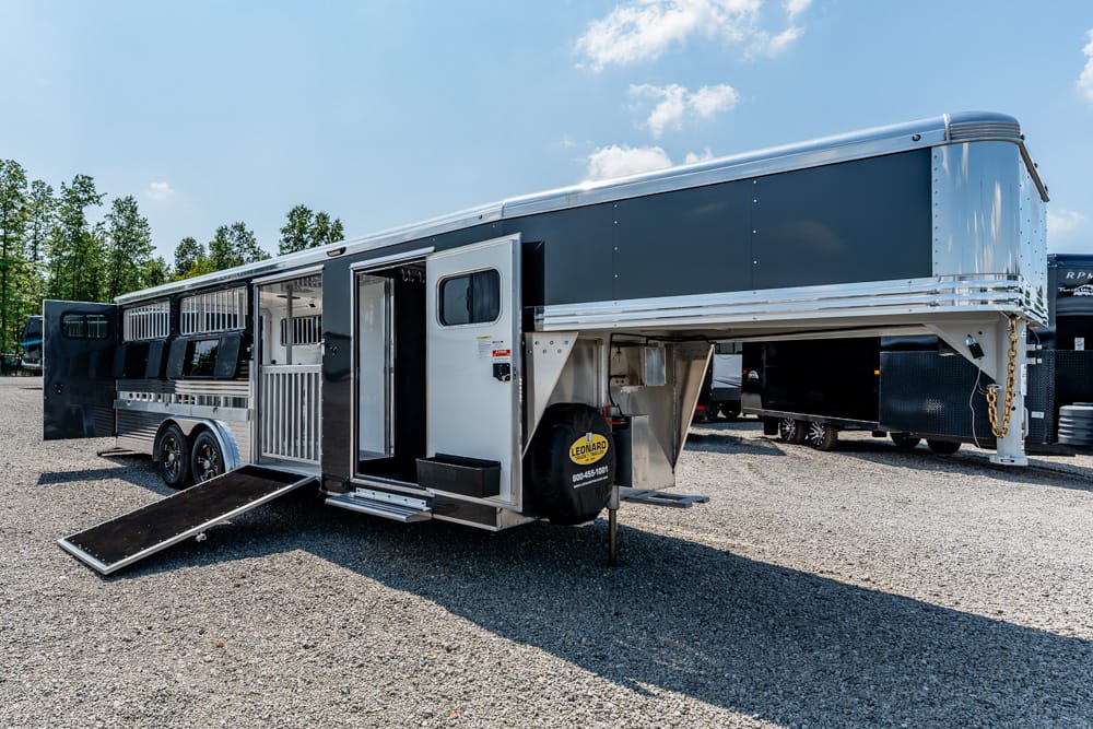 Gooseneck horse trailer with living quarters, silver/black exterior, ramp lowered, parked on a gravel lot with trees in the background.