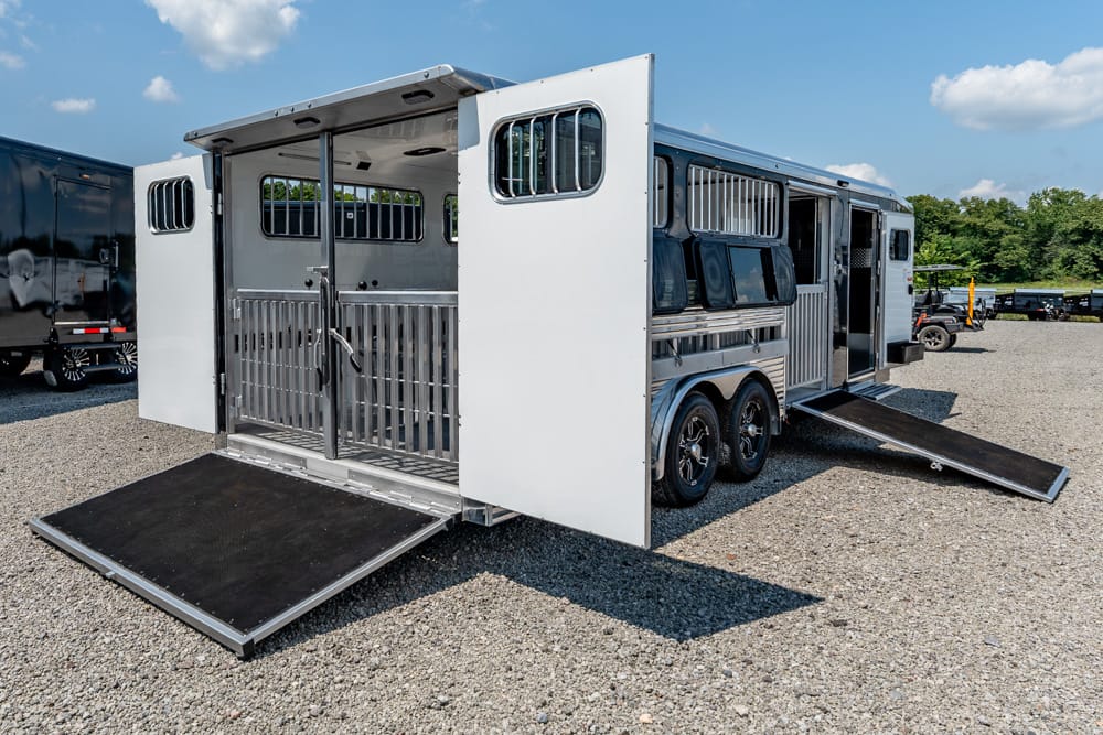 Silver horse trailer with open rear and side ramp down for loading, interior divider bars visible, parked on gravel.