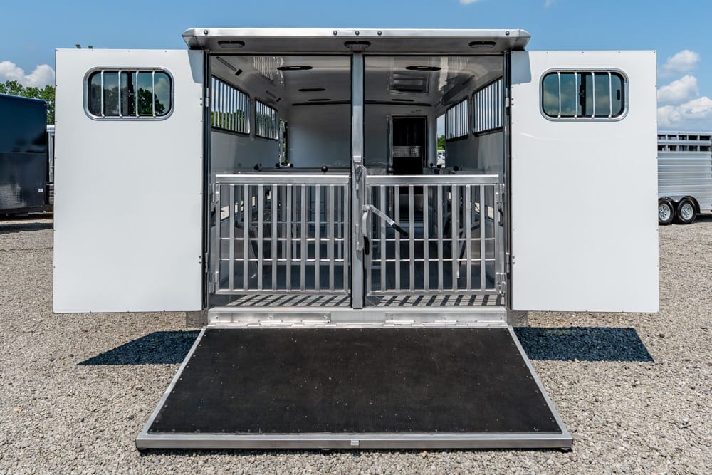 Open horse trailer with ramp lowered, showing stalls and metal dividers inside on a gravel lot.