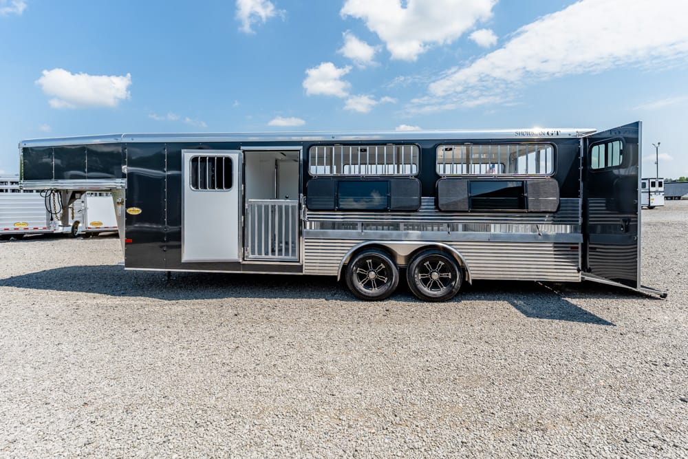 Long black horse trailer with silver lower panels and an open side door on a gravel lot under a blue sky with clouds.