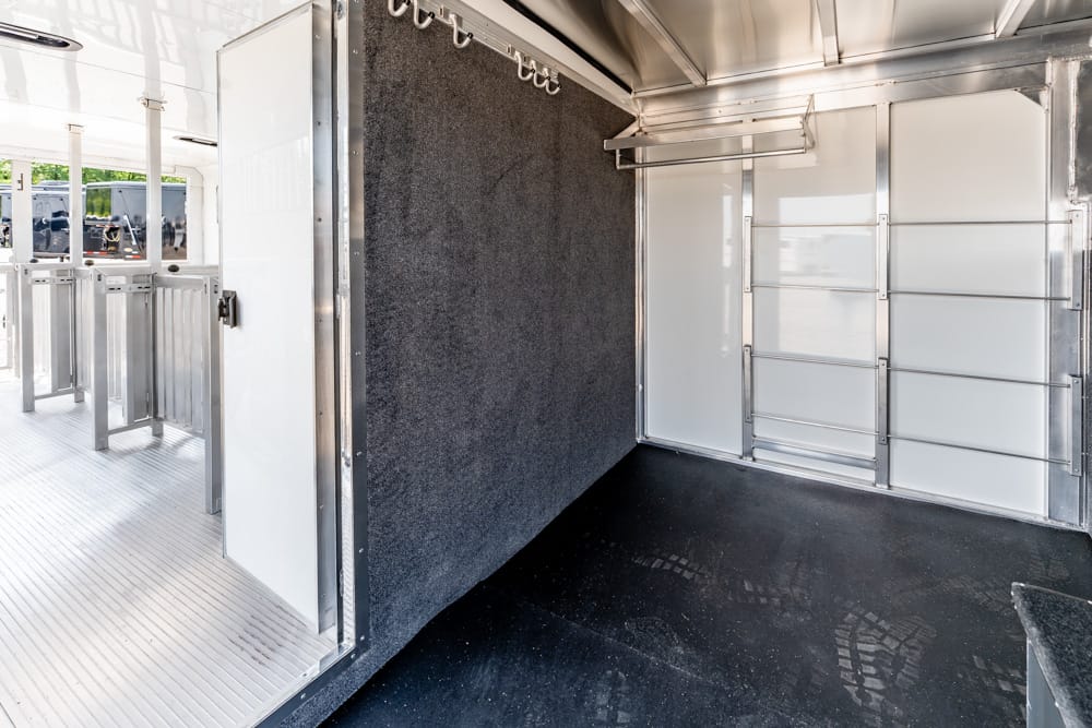 Interior of a metallic horse trailer with a dark partition wall, overhead hooks, and white panel walls on the right; black rubber floor.
