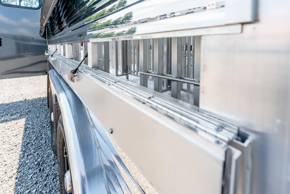 Side view of a shiny aluminum utility trailer with a built-in ladder rack and wheel on gravel.