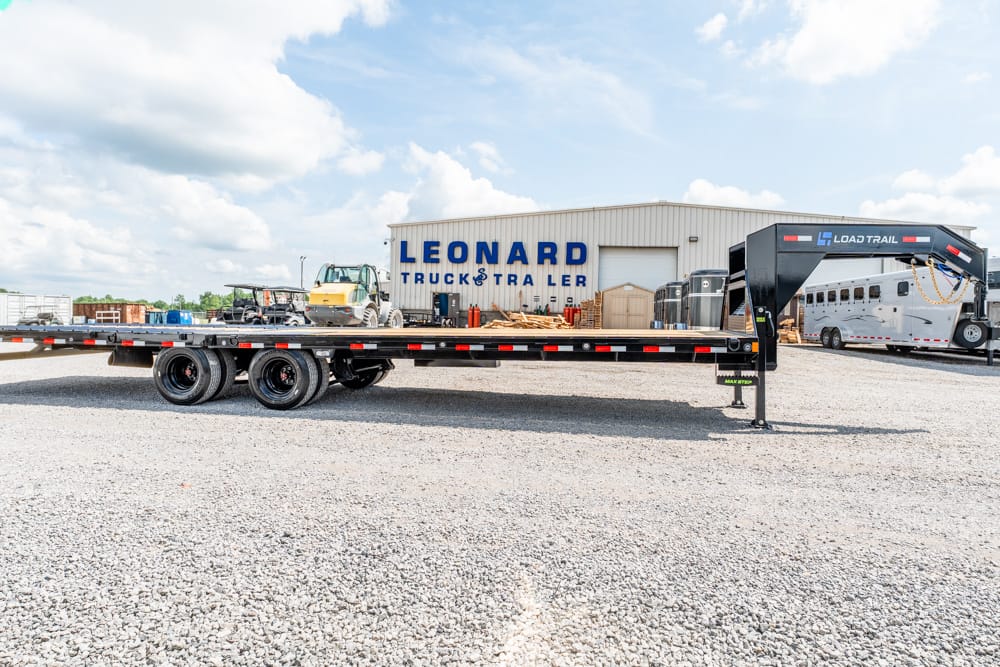 Long flatbed trailer parked in a gravel lot with a Leonard Truck & Trailer building in the background.