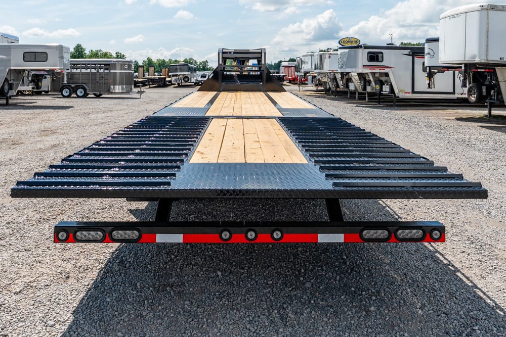 Long flatbed trailer with wooden deck and metal ramp extensions in a trailer yard, viewed from the rear edge.
