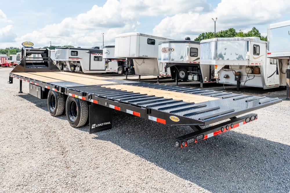 Open flatbed trailer with wooden deck in a gravel lot, surrounded by parked RVs and trailers outside a dealership.”,
