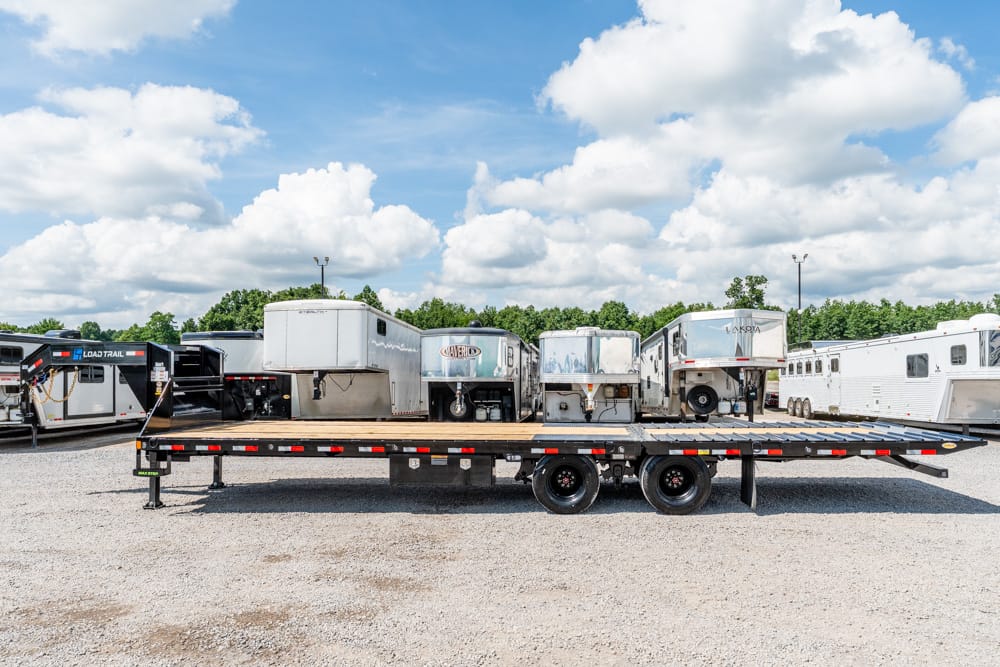 Flatbed trailer in a gravel lot with several white horse trailers parked in the background under a blue sky with clouds.