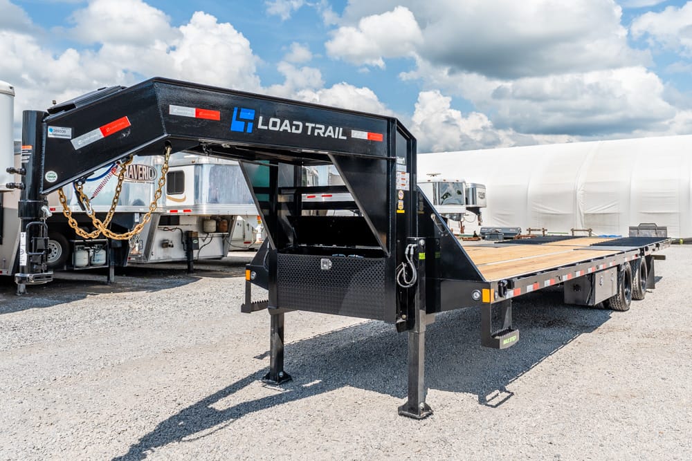 Black Load Trail gooseneck trailer with a wooden flatbed parked on gravel, chains hanging from the hitch and dual rear axles visible.