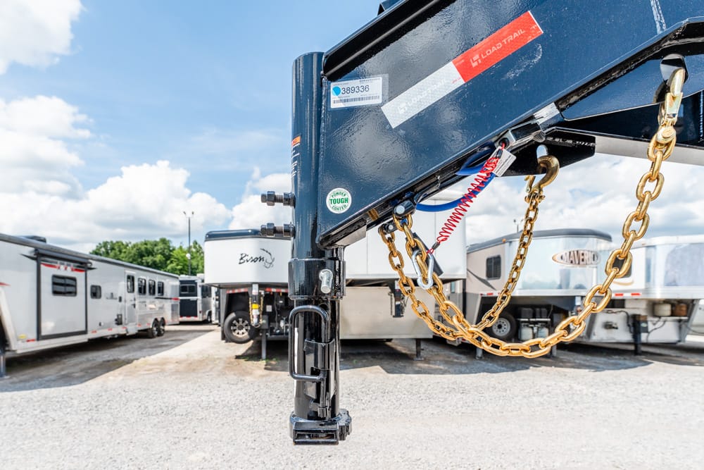 Close-up of a black gooseneck trailer hitch with gold chains looped around, in a parking lot of RV trailers under a blue sky.