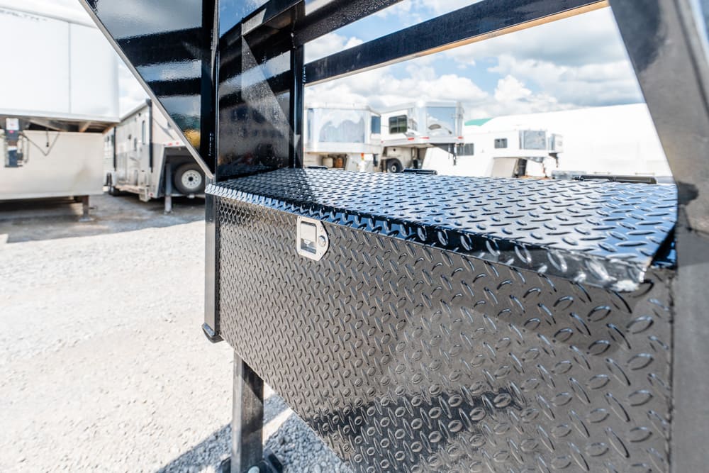 Close-up of a black diamond-plate toolbox mounted under a trailer, with other trailers in a gravel lot in the background area.