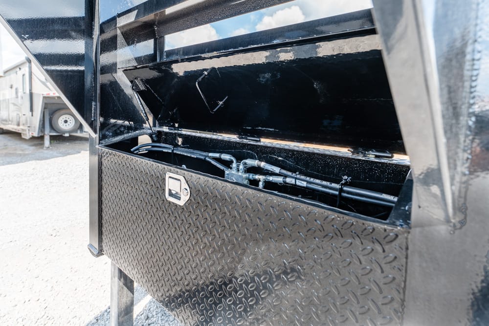 Open diamond-plate toolbox on a black dump trailer, showing hydraulic hoses inside and a latch on the front plate.