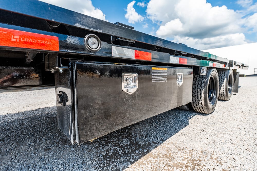 Black flatbed trailer with a side storage toolbox, dual tires, and reflective tape on a gravel lot under a blue sky with clouds.