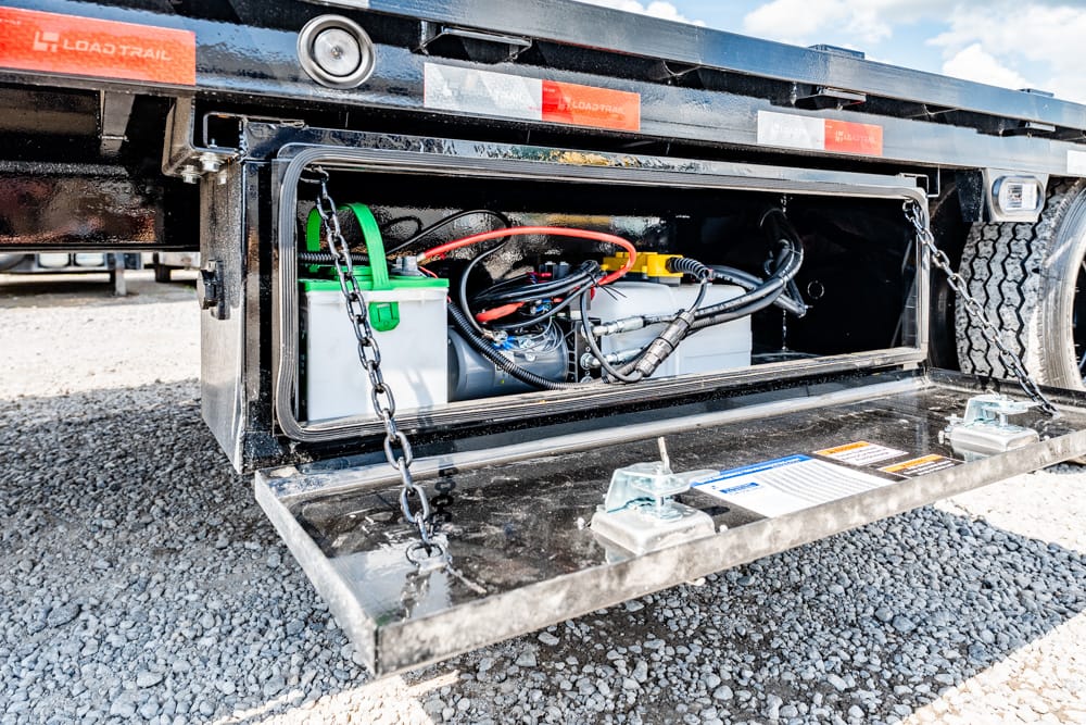 Open storage compartment of a flatbed trailer showing two batteries, cables, and wiring.