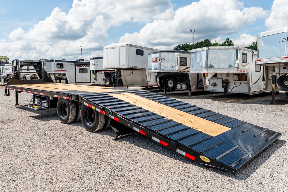 Long black flatbed trailer with a wooden ramp, parked on gravel beside several RV trailers and horse trailers in a lot.