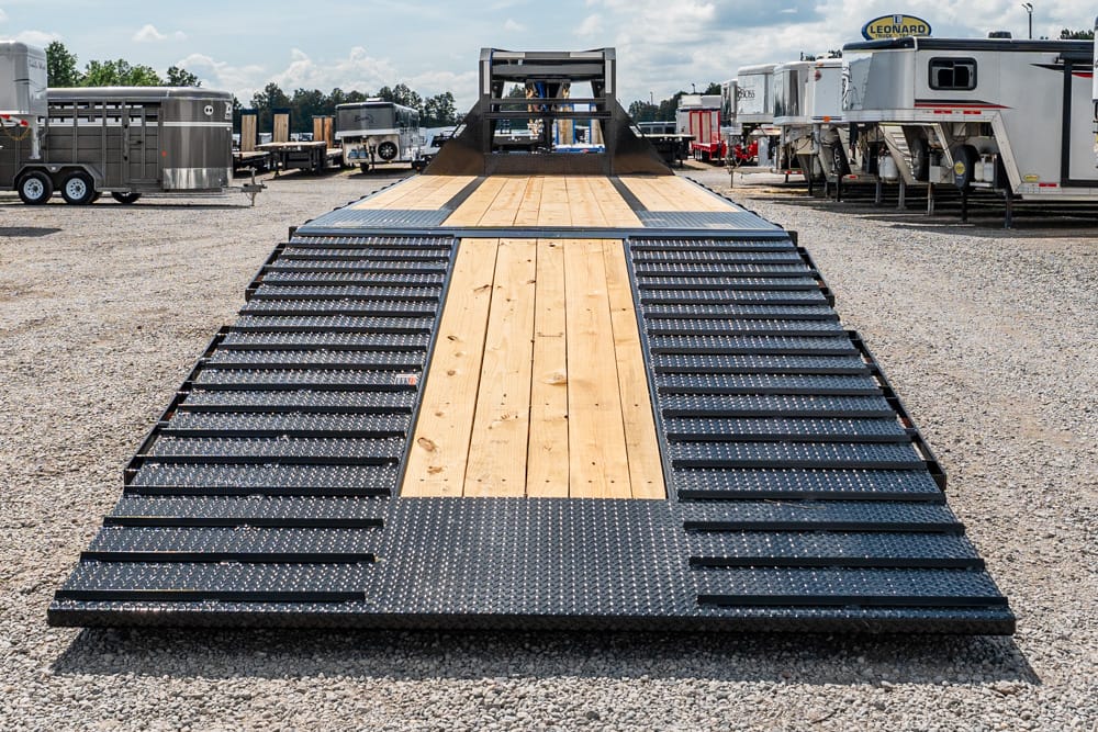 Flatbed trailer with a wood center deck and black perforated loading ramps on both sides, set in a gravel trailer lot.