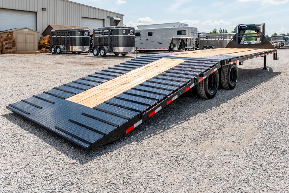 Long black flatbed trailer with a wood center deck and diamond-plate ramps extended, parked on a gravel lot with horse trailers nearby.