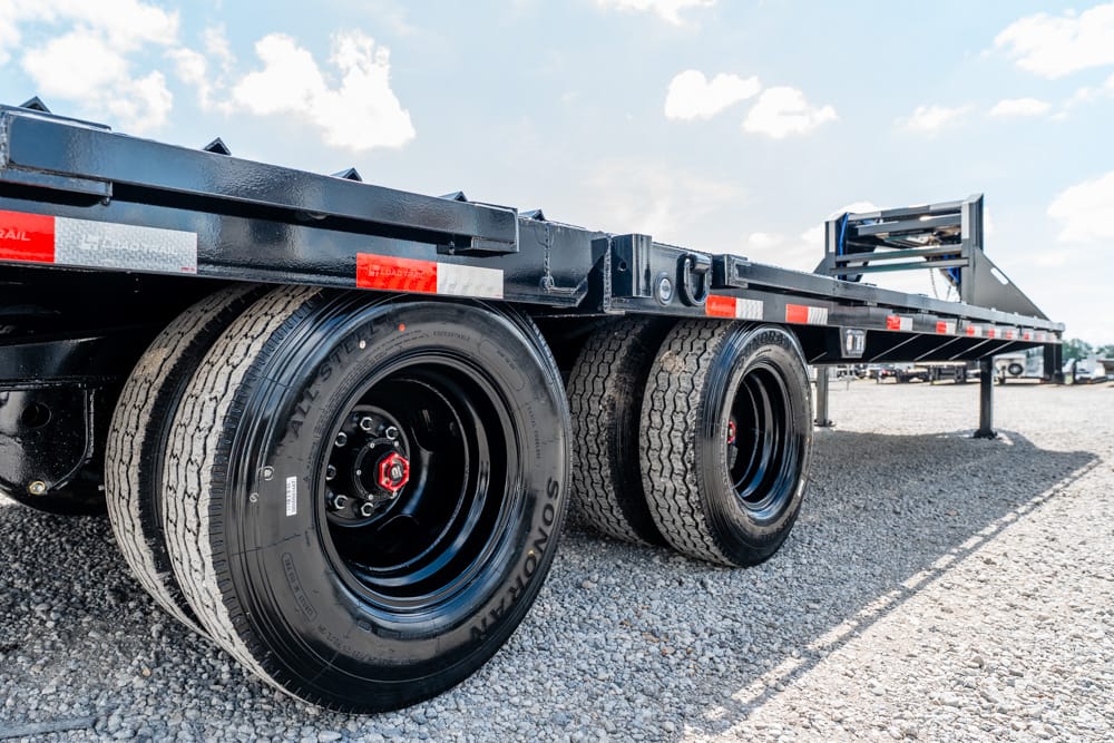 Rear view of a heavy-duty flatbed trailer showing dual rear axles with black rims on a gravel lot under a sunny sky.