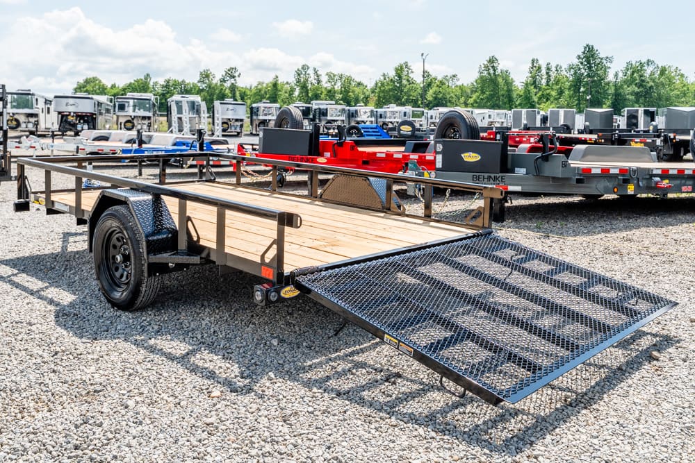 Flatbed trailer with a wooden deck and a fold-down steel loading ramp parked on a gravel lot, surrounded by other trailers.