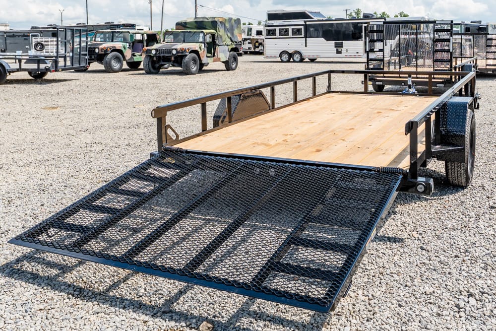 Flatbed utility trailer with a lowered mesh ramp and wooden deck in a gravel lot, ready for loading vehicles.
